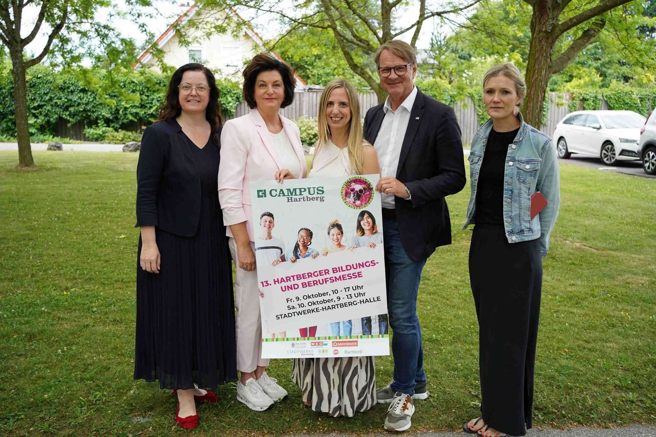 Five individuals stand on a grassy lawn, holding a large banner. The banner features images of children and text about an educational event. The individuals are dressed in formal attire, and the setting includes a house and trees in the background.