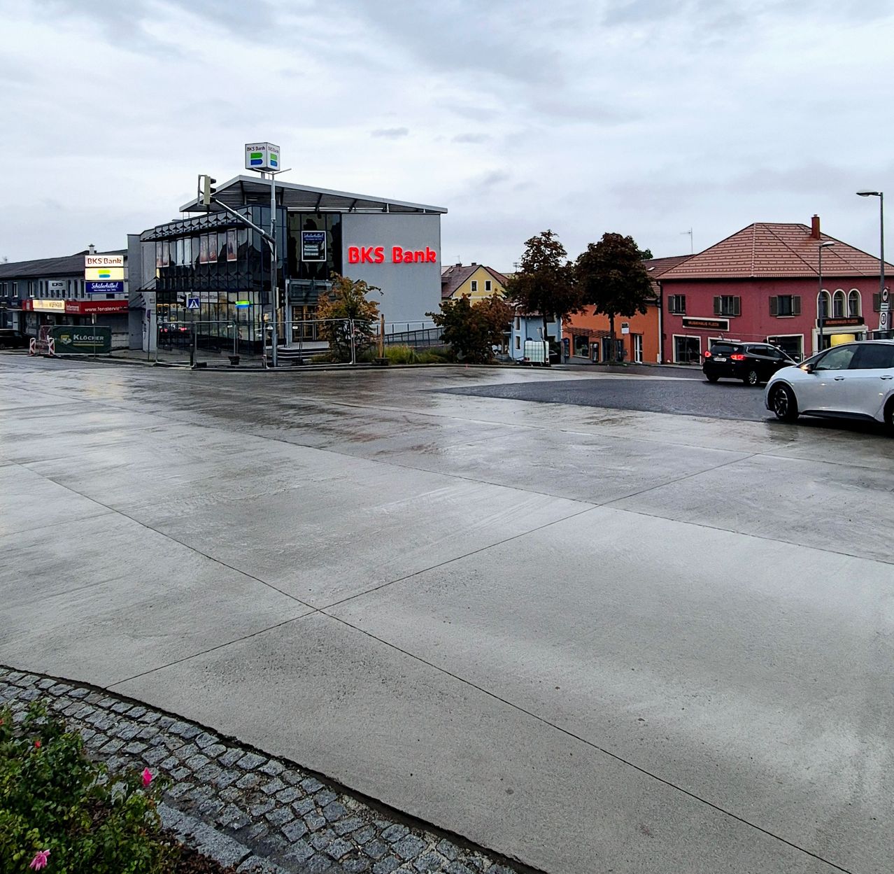 A wet street in front of a BK Bank building with a car parked nearby.