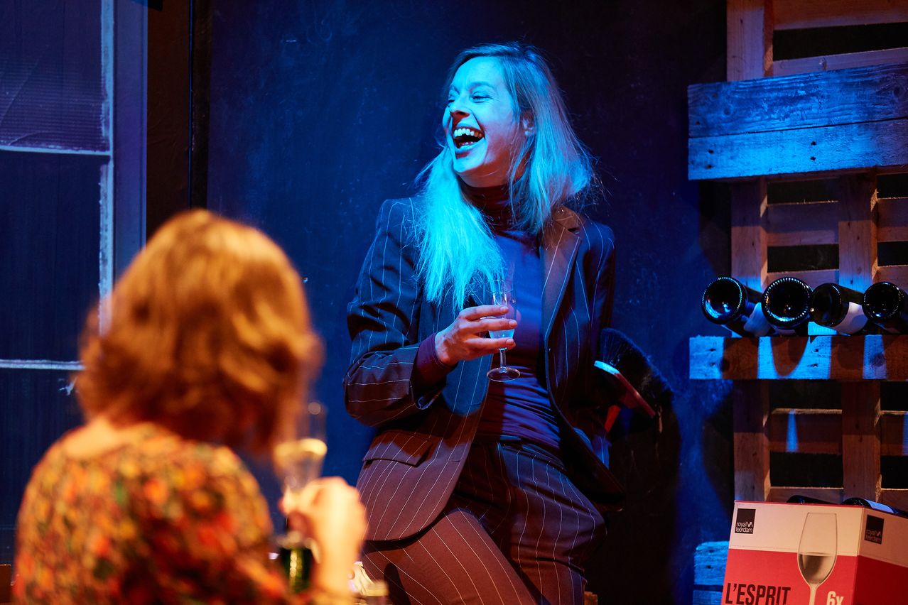A woman in a suit laughs while holding a wine glass, seated on a stage with a wooden backdrop. Another woman holds a glass in the foreground.