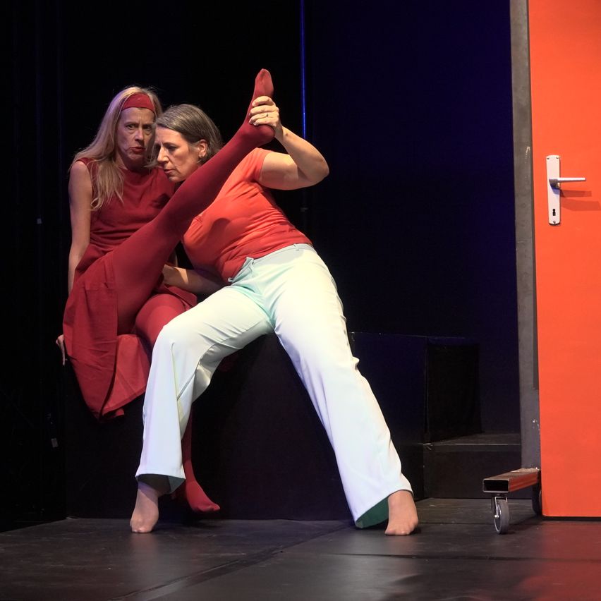 Two women on stage, one in red and one in white, performing a dance routine. The woman in white is stretching her leg while the other watches.