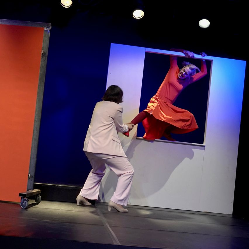 A woman in a pink suit helps another woman in a red dress climb out of a framed picture on a stage.