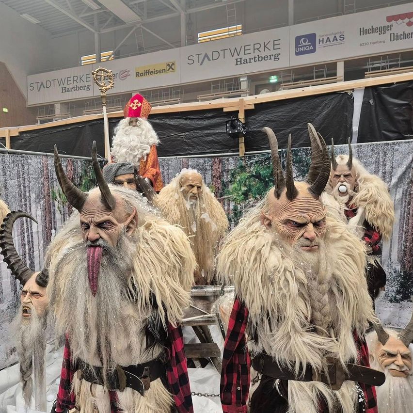 A group of people dressed in horned costumes, possibly for a festival or event. They are standing in a row, with one person wearing a Santa Claus costume. Behind them, there is a wall with a banner and a logo.