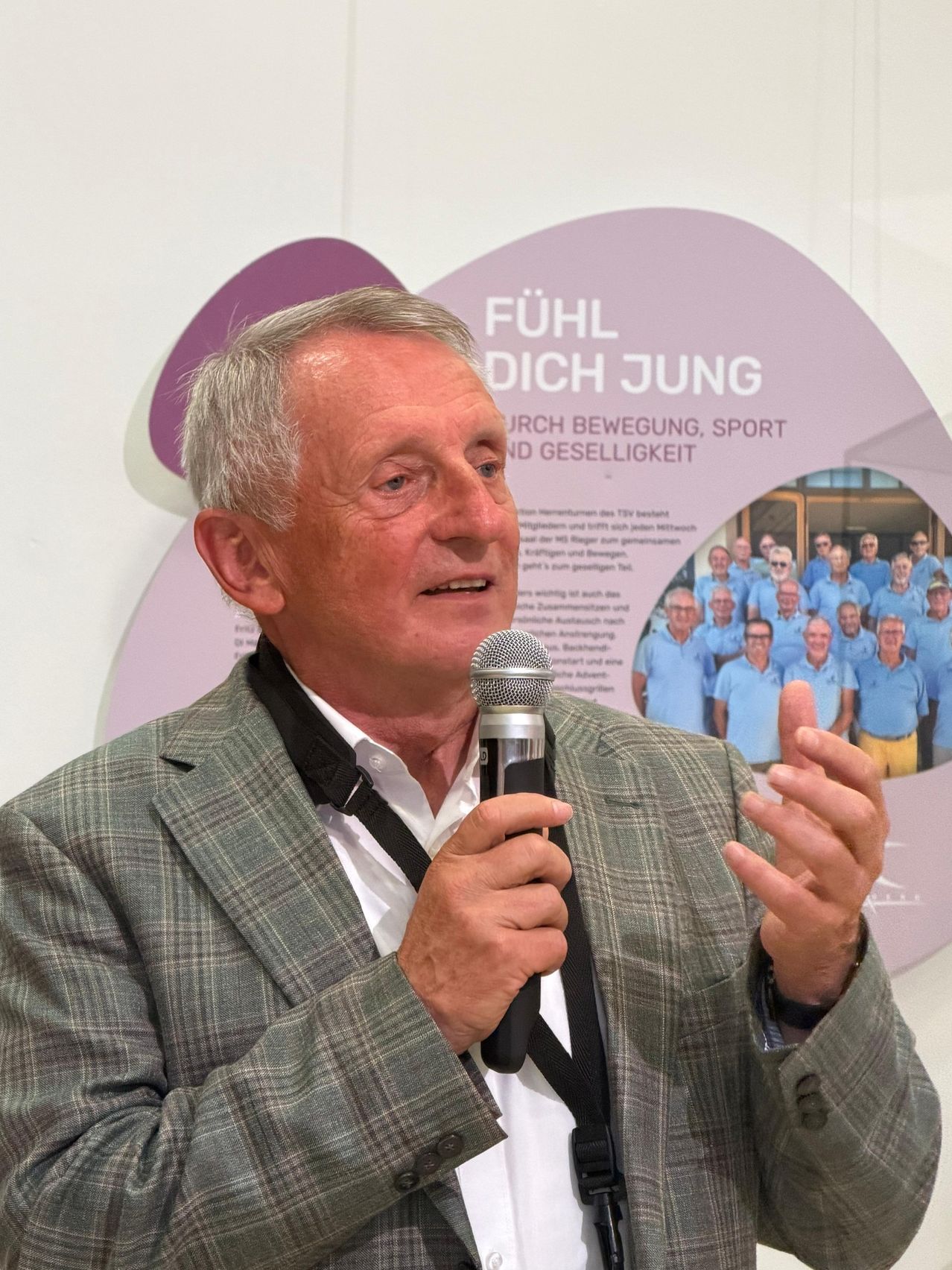 Ein Mann in einem karierten Anzug spricht in ein Mikrofon bei einer öffentlichen Veranstaltung. Dahinter befindet sich ein Hintergrund mit dem Text Fühl dich Jung, umgeben von Bildern von Menschen.