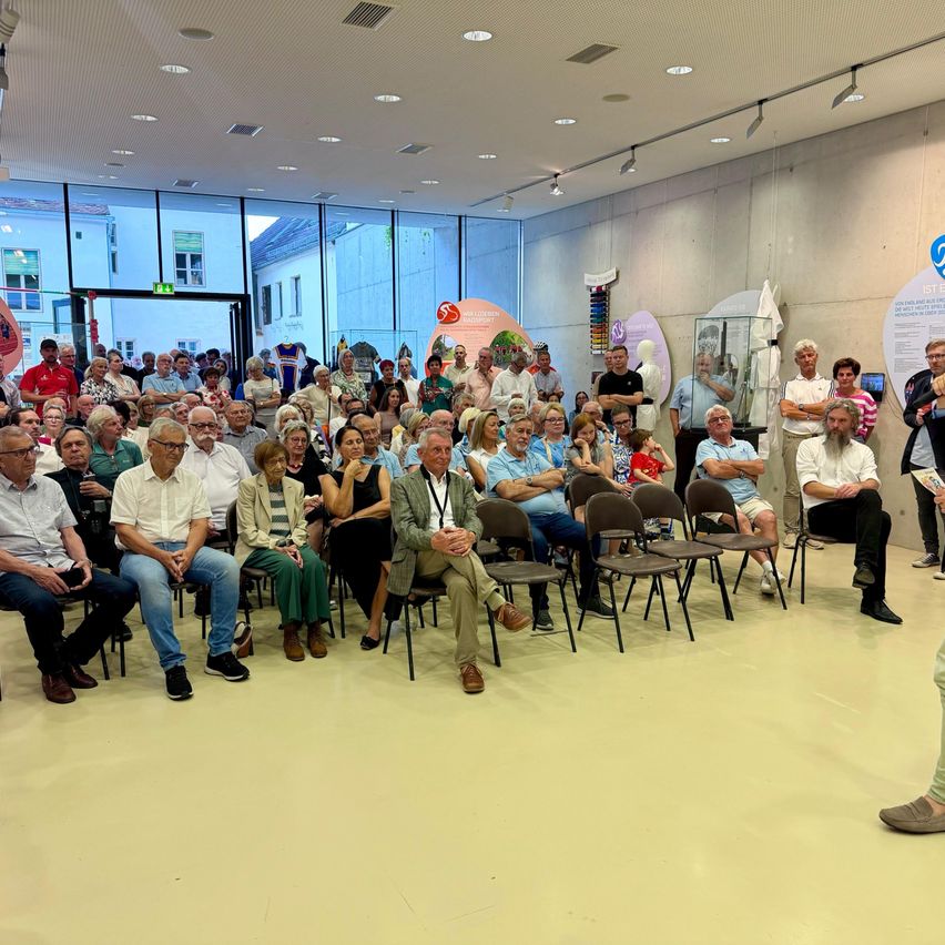Eine große Menschenmenge sitzt in Reihen in einem Konferenzsaal und schaut nach vorne. Ein Mann vorne steht und spricht.