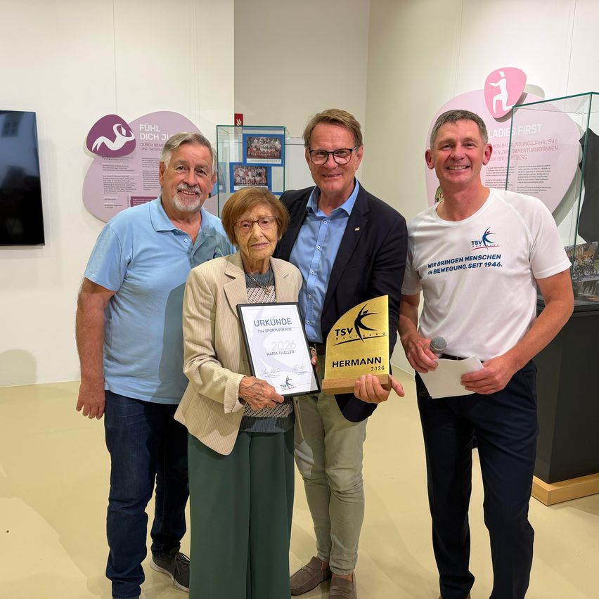 Vier Personen stehen gemeinsam und halten Auszeichnungen, lächeln für ein Foto. Eine Frau hält ein gerahmtes Zertifikat und eine Auszeichnungsplakette, während die anderen Mikrofone halten.