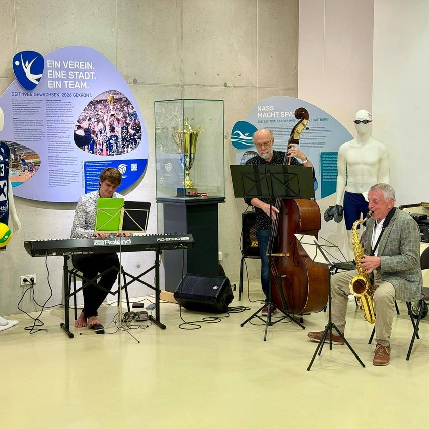 Vier Musiker spielen drinnen mit einem Pokal, einer Puppe und Informationstafeln im Hintergrund.