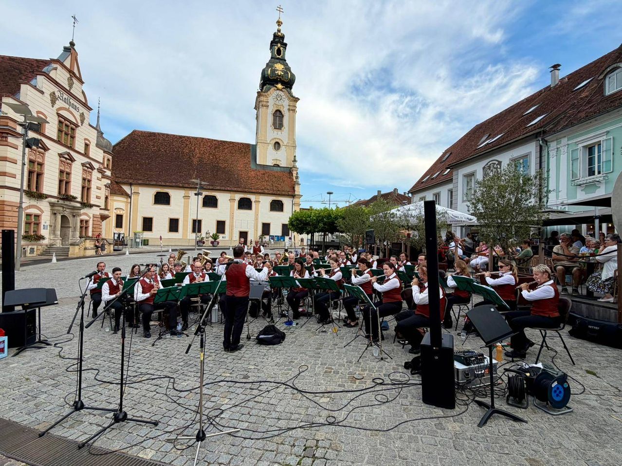 Ein Orchester spielt auf einer Kopfsteinpflasterstraße vor einem großen Gebäude mit Turm und braunem Dach.