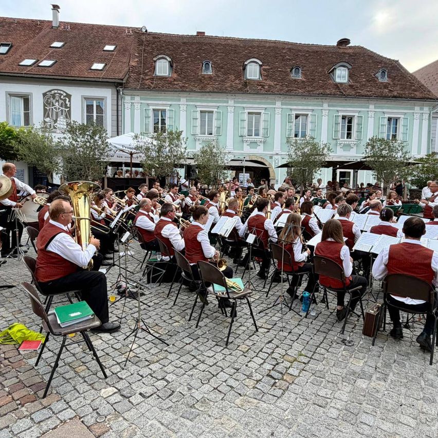 Ein Blasorchester spielt im Freien vor einem historischen Gebäude. Musiker sitzen auf Stühlen, halten Instrumente, ein kleines Publikum ist anwesend.