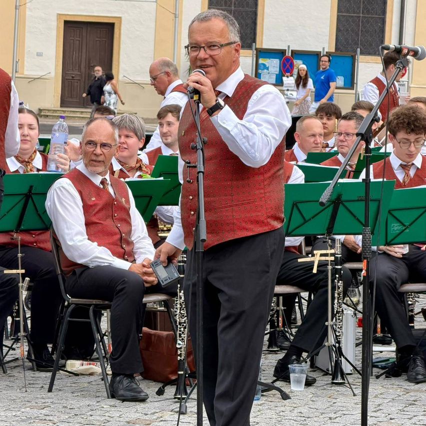 Eine Band probet in einem offenen Bereich, mit Musikern, die vor grünen Ständern sitzen. Ein Mann in einem roten West steht und hält ein Mikrofon.