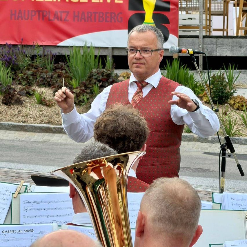 Ein Mann mit Brille dirigiert eine Band auf einer Bühne. Hinter ihm steht ein Mikrofon auf einem Ständer. Die Bandmitglieder spielen ihre Instrumente. Ein Banner mit dem Text 'Hauptplatz Hartberg' ist hinter ihnen sichtbar.