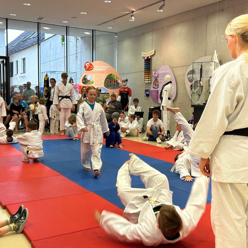 Kinder führen Karate-Bewegungen in einem Studio mit Zuschauern durch. Ein Kind fällt auf die Matte.