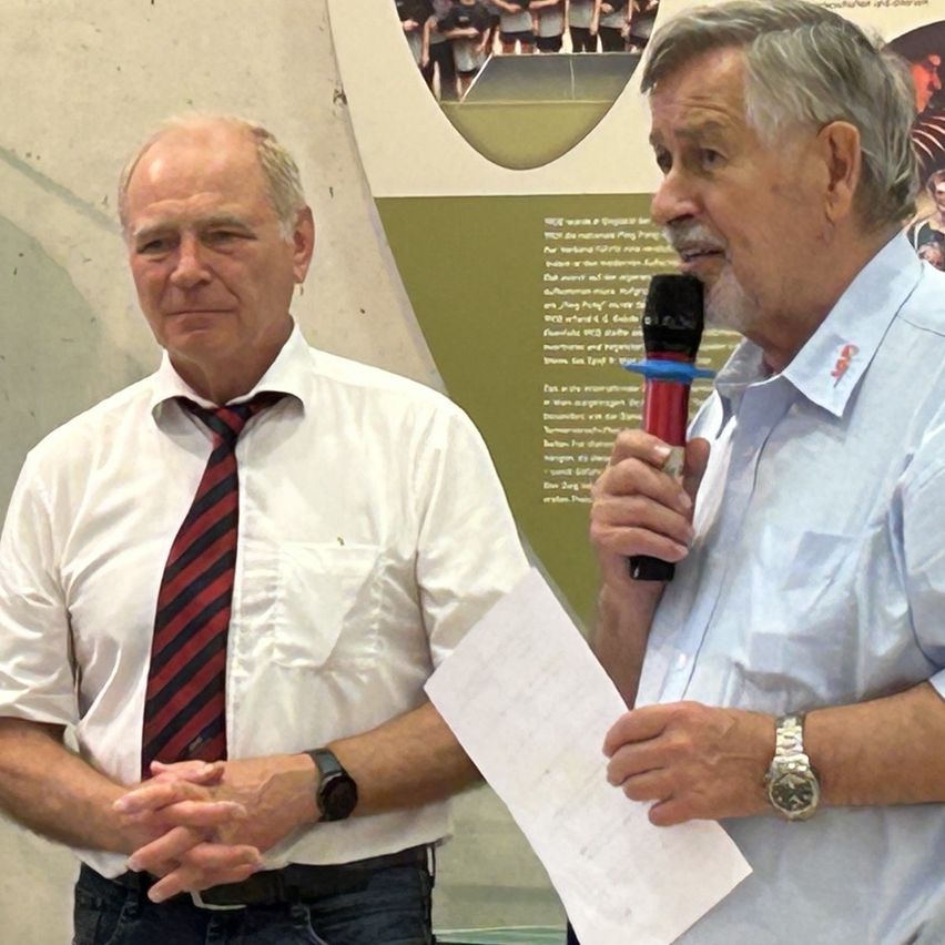 Zwei Männer in formeller Kleidung stehen vor einer Wand. Ein Mann hält ein Mikrofon und scheint zu sprechen. Der andere Mann trägt eine Krawatte und hört aufmerksam zu.