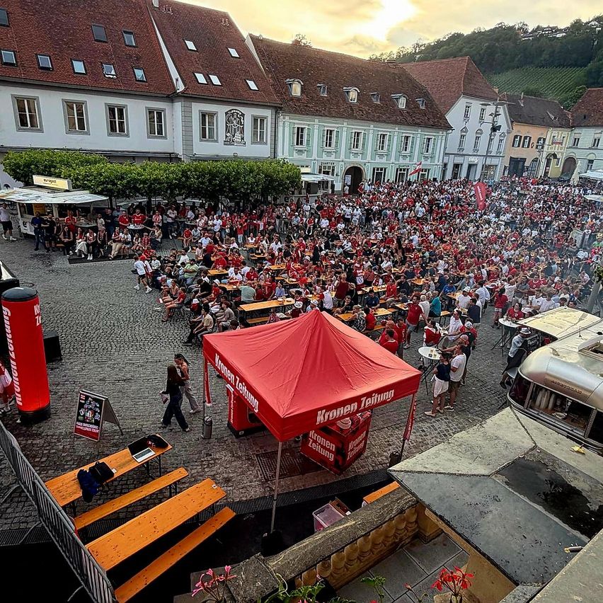 Eine Luftaufnahme einer großen Menschenmenge in einem Freiluftbereich. Viele Menschen sitzen, und ein rotes Zelt mit dem Text 'Kronen' befindet sich im Vordergrund. Gebäude und Grünflächen umgeben das Gebiet.