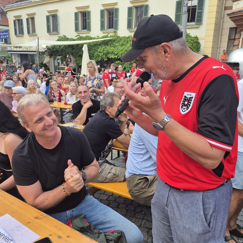 Ein Mann in einem roten Fußballtrikot spricht in ein Mikrofon und spricht eine Menschenmenge in einem Biergarten an. Ein anderer Mann lächelt im Vordergrund, während er an einem Tisch sitzt.