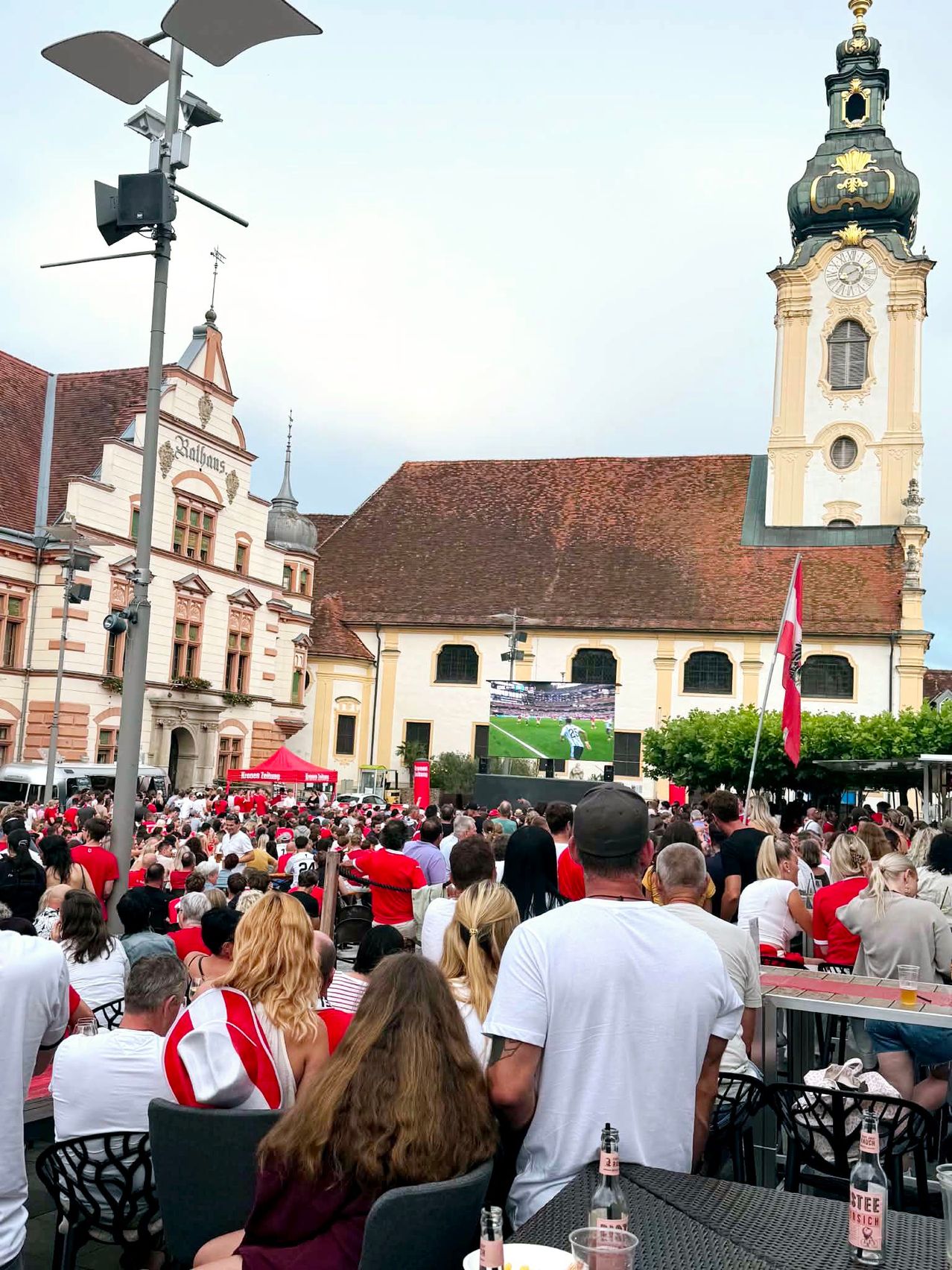 Eine Menschenmenge versammelt sich vor einem großen Gebäude mit einem Turm und einer großen Leinwand, die ein Fußballspiel zeigt. Einige Menschen tragen rote T-Shirts.