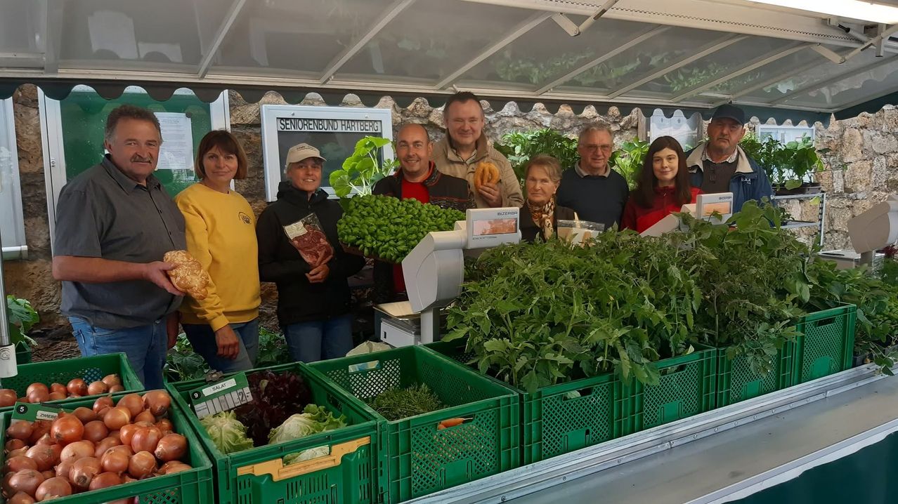 Eine Gruppe von Menschen ist auf einem Bauernmarkt, lächelt und posiert für ein Foto. Sie sind von grünen Kisten mit Gemüse und einer Waage umgeben. Ein Schild steht Seniorenbund Hartberg.