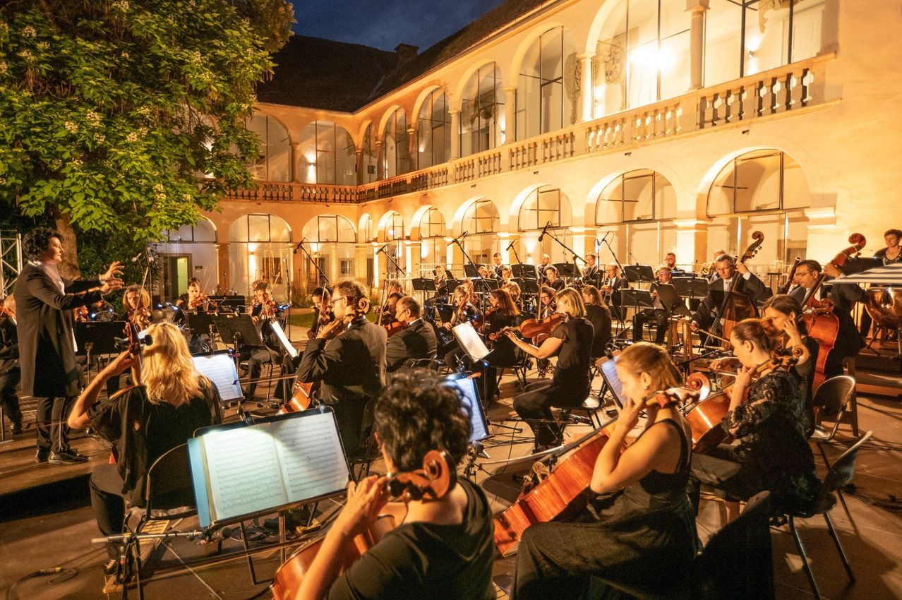 Ein Orchester spielt bei Nacht in einem Innenhof, beleuchtet von Lampen. Musiker spielen Violinen, Celli und andere Instrumente. Die Architektur ist historisch mit Bogenfenstern.