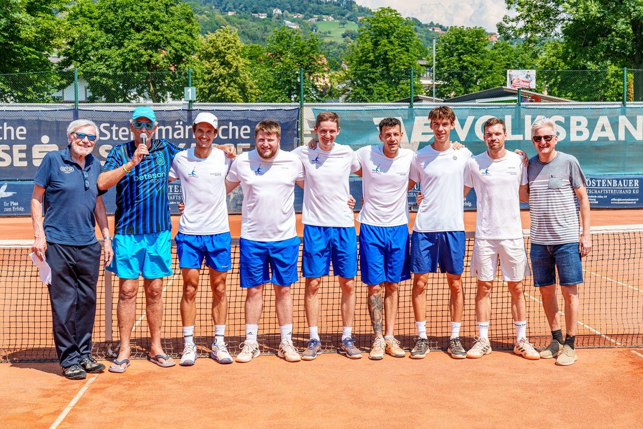 Sechs Männer posieren vor einem Tennisnetz für ein Foto. Sie tragen alle ähnliche weiße und blaue Outfits. Ein Mann in einem gestreiften Hemd hält ein Mikrofon. Hinter ihnen ist ein grüner Zaun mit Bannern.
