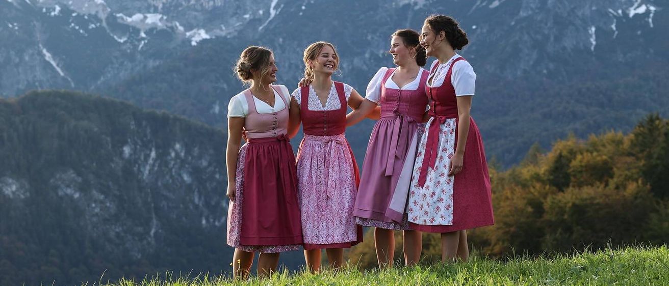 Vier Frauen in traditionellen bayerischen Kleidern stehen lächelnd auf einem Grashügel, mit verschneiten Bergen im Hintergrund.