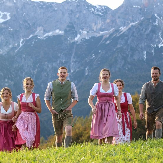 Eine Gruppe lächelnder junger Leute steht auf einer Grasfläche mit Bergpanorama im Hintergrund. Sie tragen traditionelle Kleidung. Das Wetter scheint sonnig zu sein.