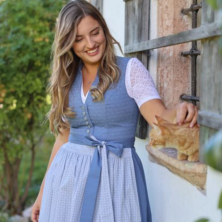 Eine Frau mit langen blonden Haaren trägt ein blaues Kleid mit weißen Spitzendetails und einem Gürtel, lächelt und hält eine kleine Katze auf der Fensterbank.