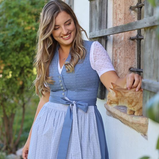 Eine Frau mit langen blonden Haaren trägt ein blaues Kleid mit weißen Spitzendetails und einem Gürtel, lächelt und hält eine kleine Katze auf der Fensterbank.