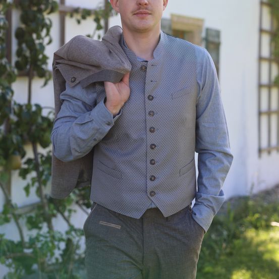 Ein Mann steht vor einem Gebäude, hält einen grauen Mantel und trägt eine gemusterte Weste mit mehreren Knöpfen. Er hat einen ernsten Gesichtsausdruck und ist in graue Hosen gekleidet.