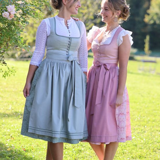 Zwei Frauen in traditionellen bayerischen Dirndln stehen auf einer Wiese und lächeln für ein Foto.
