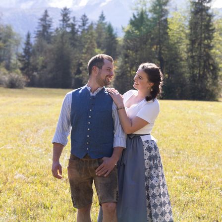 Ein Paar steht in einem Grasfeld, lächelt und sieht sich an. Der Mann trägt eine blaue Weste und braune Shorts, während die Frau ein weißes Oberteil und einen gemusterten Rock trägt. Dahinter sind Bäume und Berge.