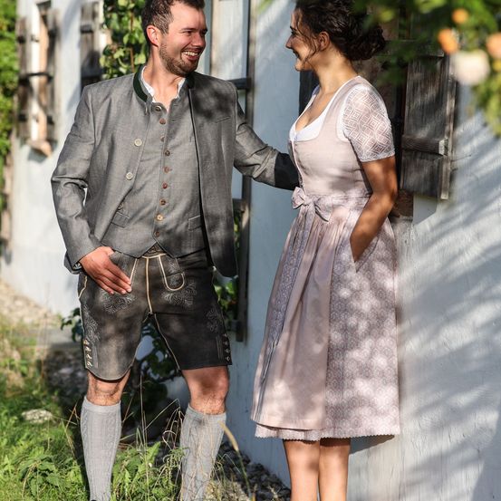 Ein Mann und eine Frau in traditioneller Kleidung stehen eng beieinander, lächeln und schauen in eine Richtung. Der Mann trägt einen grauen Anzug und schwarze Shorts, während die Frau ein rosa Kleid trägt.
