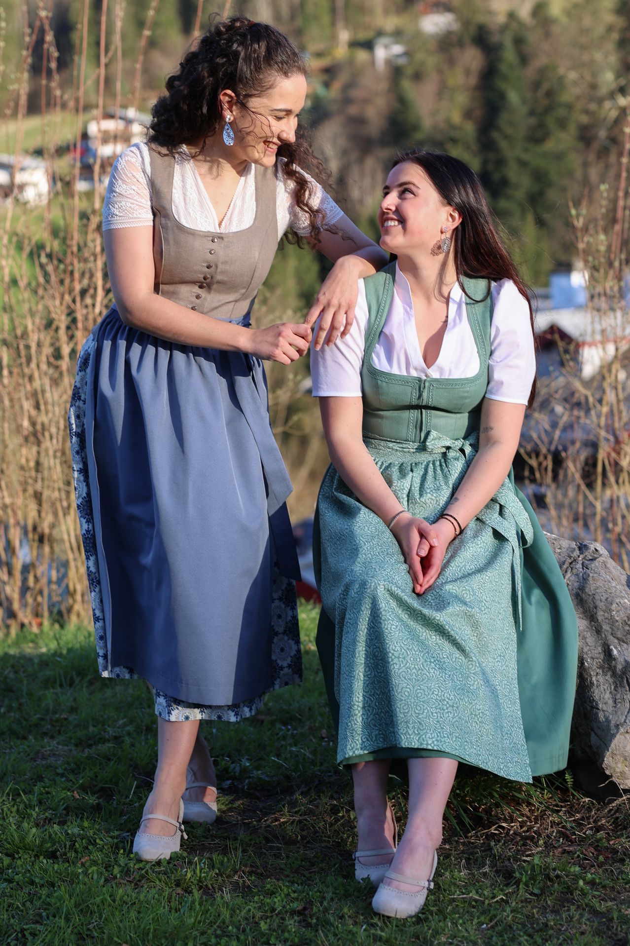 Zwei Frauen in traditioneller Kleidung stehen auf einem Rasen, eine lächelt und die andere hat die Hände gefaltet. Die Frau auf der linken Seite trägt ein hellblaues Kleid mit Blumenmuster. Die Frau auf der rechten Seite trägt ein grünes Kleid. Sie sind von kahlen Ästen umgeben.