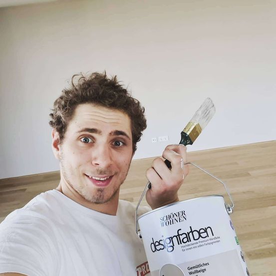 Ein Mann hält einen Pinsel und eine Farbeimer mit der Aufschrift 'designfarben'. Er lächelt, möglicherweise in einem Haus, das renoviert wird.