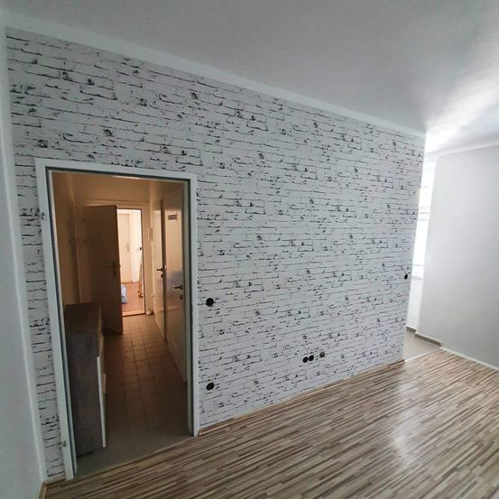 Ein leerer Raum mit einer weißen Ziegelwand, Holzboden und offenen Türen, die zu einem anderen Zimmer mit gefliestem Boden führen.