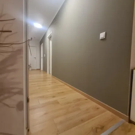 Ein leerer Korridor mit Holzböden und grauen Wänden. An der Wand befindet sich ein weißer Schalter. Der Korridor führt zu einem Raum mit einer weißen Tür.