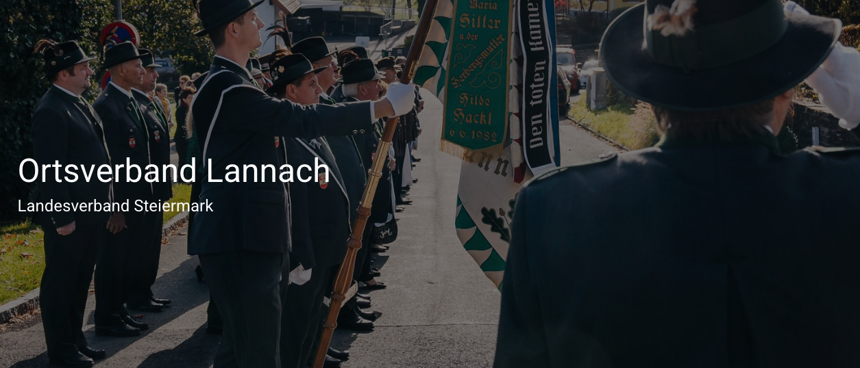 Eine Gruppe von Personen in Uniform, die Fahnen halten, steht auf einer Straße mit dem Wort 'Lannach' oben.