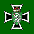 Das Bild zeigt ein Wappen auf grünem Hintergrund mit einem weißen Löwen, der eine rote Zunge und eine rote Krone hat. Der Löwe ist von einem grünen Schild umgeben, der wiederum von einem weißen Kreuz umkreist wird.