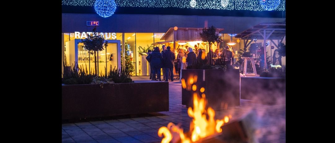 Eine Menschenmenge steht vor einem Gebäude mit leuchtenden Lichtern und einem Feuerkorb, unter einem Himmel mit festlichen Lichtern.