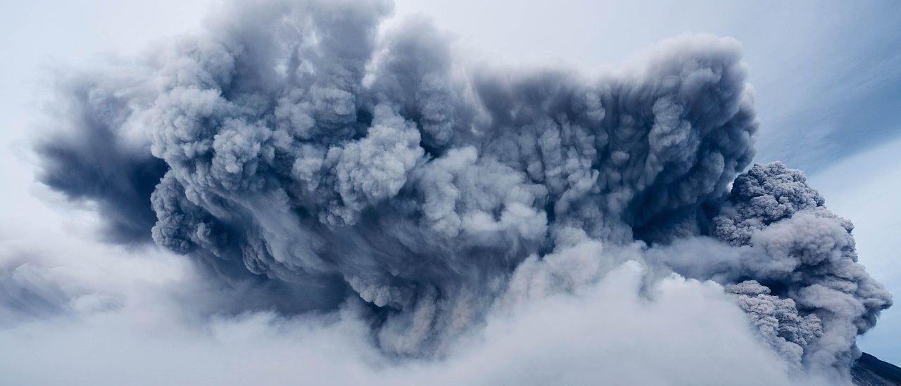 Eine große, dichte Rauch- und Aschewolke steigt von einem Vulkan gegen einen grauen Himmel auf. Der Rauch steigt aufwärts und verdeckt den Himmel. Unten ist eine Landschaft mit Bäumen und Bergen zu sehen.