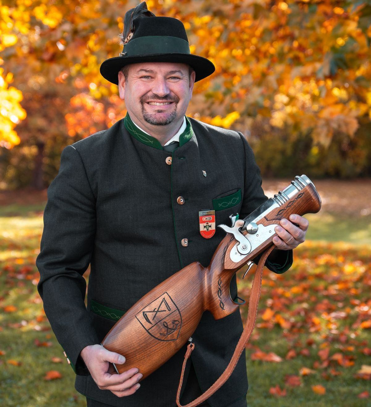 Ein Mann in traditioneller Kleidung hält eine Holzwaffe und lächelt im Freien, mit Herbstlaub im Hintergrund.