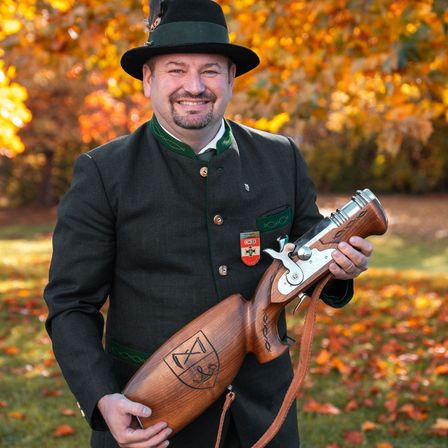 Ein Mann in traditioneller Kleidung hält eine Holzwaffe und lächelt im Freien, mit Herbstlaub im Hintergrund.