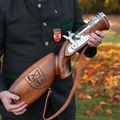 Ein Mann in traditioneller deutscher Kleidung hält ein vintage-stilvolles Gewehr und lächelt fröhlich in einer sonnigen Außenumgebung.