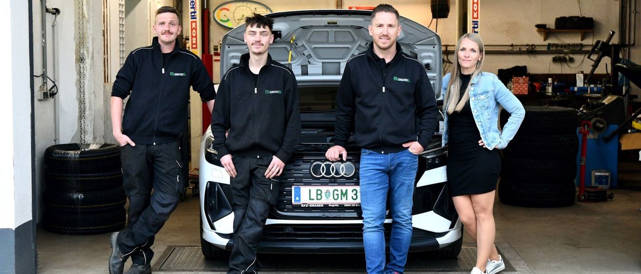 Four people are standing in front of a white Audi car in a garage, with the hood open. The man on the left is wearing a black jacket.