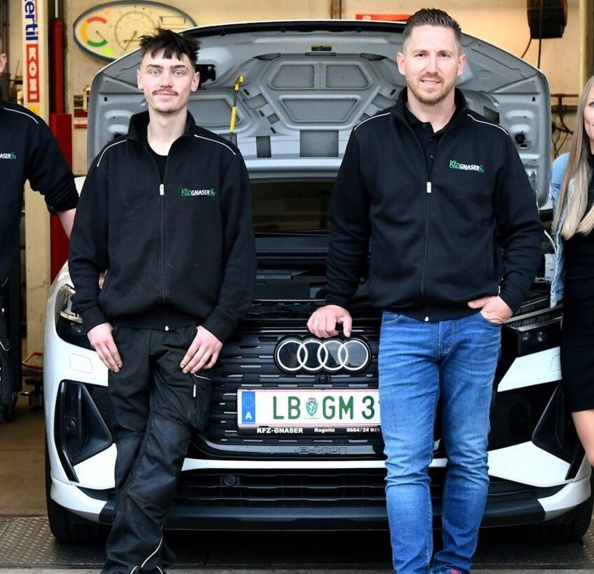 Vier Personen stehen vor einem weißen Audi-Auto in einer Garage, mit offenem Motorhaube. Der Mann auf der linken Seite trägt einen schwarzen Mantel.