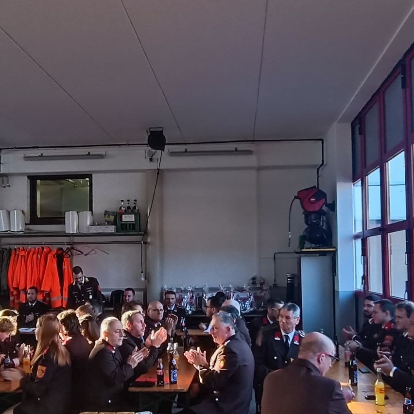 Eine Gruppe uniformierter Personen, möglicherweise Militärangehörige, versammelt sich in einem Raum. Sie klatschen und scheinen zu feiern. Flaschen und Gläser stehen auf den Tischen vor ihnen.