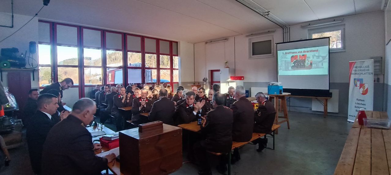 Eine Versammlung von Menschen in Anzügen findet in einem Raum mit großen Fenstern, Holzbänken und Tischen statt. Eine Person klatscht, und andere beobachten.