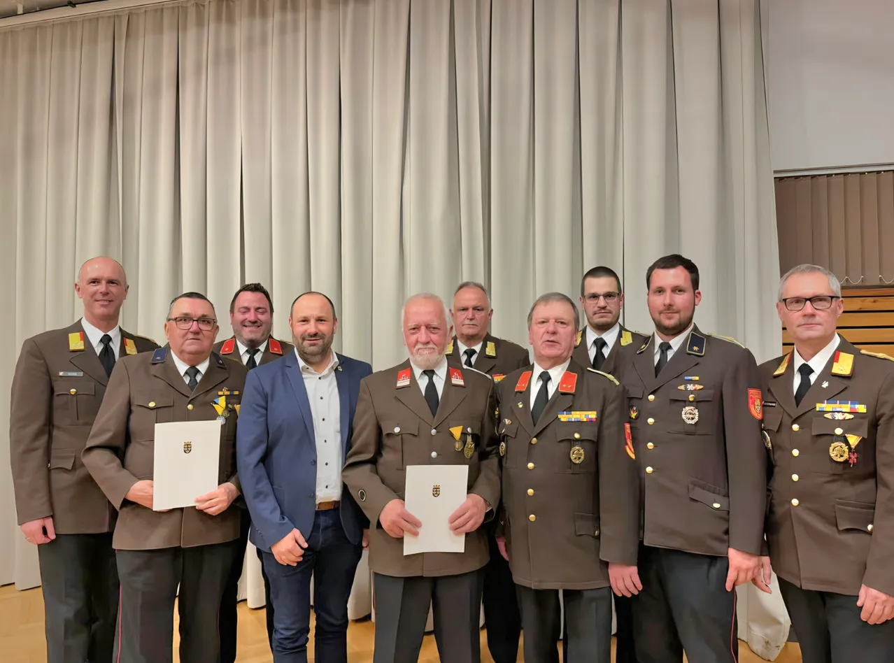 Eine Gruppe von Männern in Uniform, die Urkunden halten, steht vor einem weißen Vorhang. Ein Mann in Anzug steht unter ihnen.