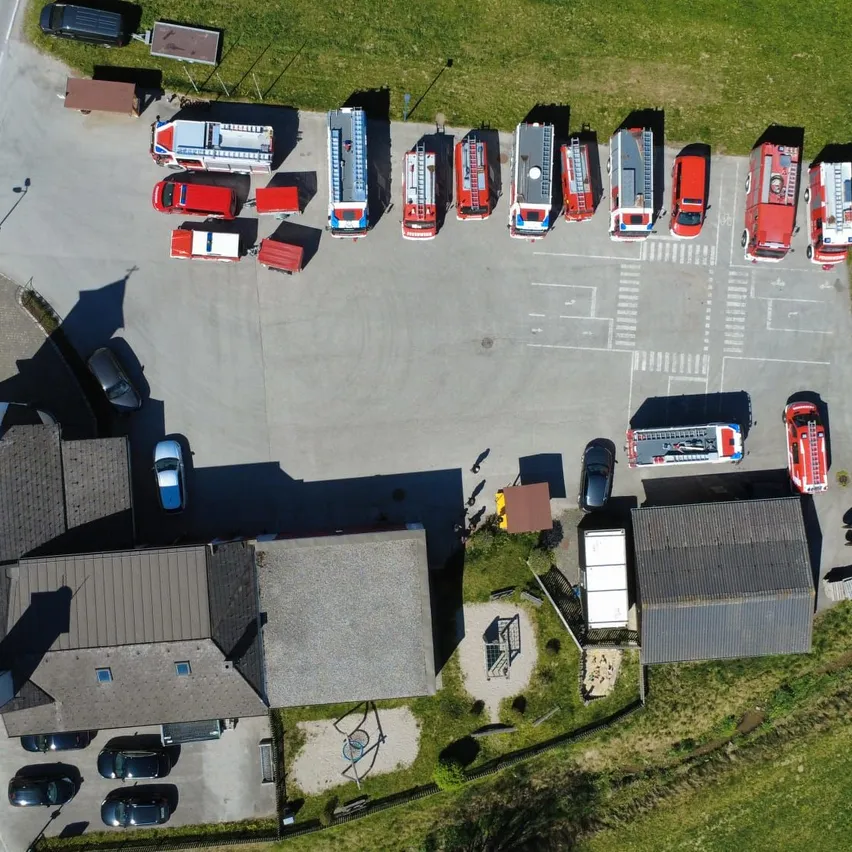Eine Vogelperspektive eines Parkplatzes mit mehreren roten und weißen Feuerwehrfahrzeugen, zusammen mit anderen Fahrzeugen. Gebäude sind an den Seiten des Parkplatzes sichtbar.