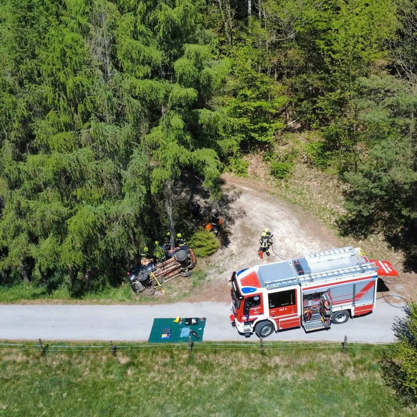 Eine Draufsicht zeigt ein umgekipptes Fahrzeug in der Nähe einer Straße in einem Wald. Feuerwehrleute kümmern sich um das Fahrzeug, einer von ihnen benutzt einen Schlauch. Ein Feuerwehrwagen ist in der Nähe geparkt.