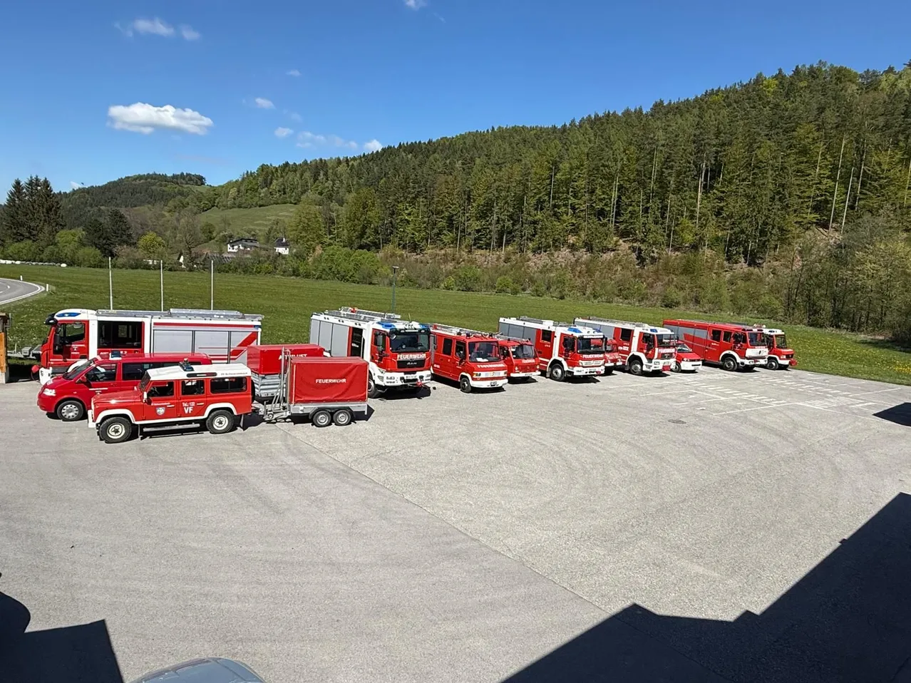 Mehrere rote Feuerwehrfahrzeuge sind in einer Reihe in einem offenen Bereich geparkt. Im Hintergrund sind Bäume und ein Haus zu sehen.