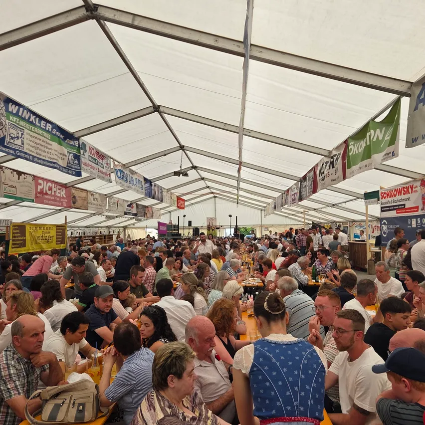 Eine große Menschenmenge versammelt sich unter einem weißen Zelt und isst an Tischen. Banner hängen von der Decke, und viele Menschen tragen Hüte und Brillen. Die Atmosphäre ist lebhaft, mit Menschen, die sich unterhalten und ihre Mahlzeiten genießen.